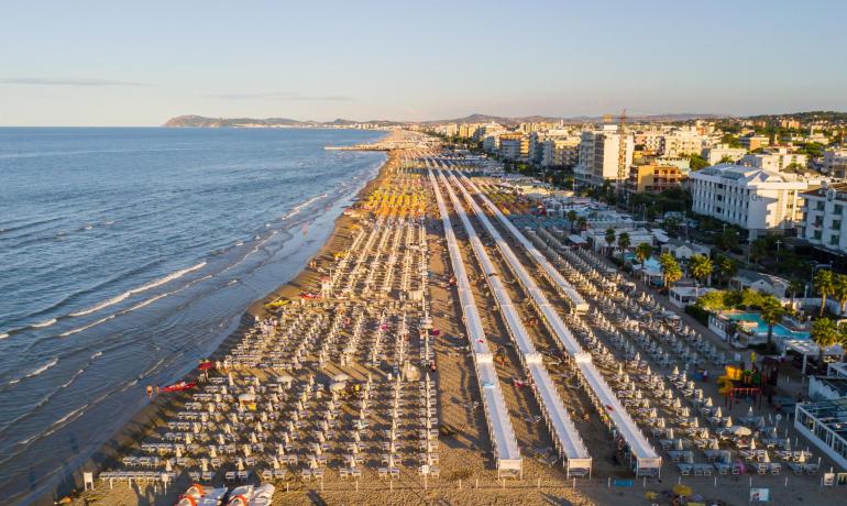Spiaggia affollata con ombrelloni e mare calmo, città sullo sfondo.