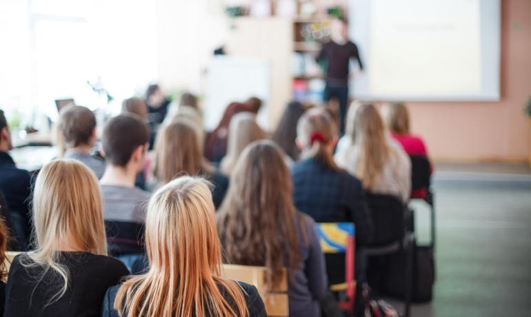 Studenti in aula ascoltano una lezione con un insegnante davanti.